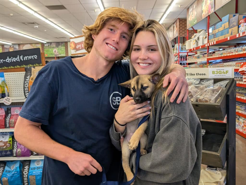 A young man and woman smile while holding a small dog in a pet store. The man wears a blue shirt and the woman is in a gray sweatshirt. Shelves stocked with pet supplies are visible in the background, indicating a moment related to pet adoption.