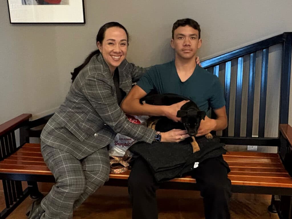 A woman and a young man are seated on a wooden bench inside a building. The woman smiles and poses with her arm around the boy, who is holding a small dog in his lap. Both are wearing casual clothes, and there are items like blankets and treats visible nearby, indicating a recent adoption.