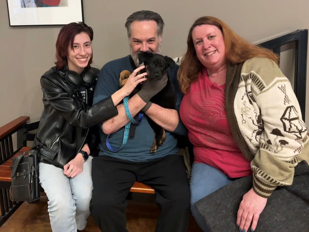 A man seated in the middle holds a small dog, while a woman on his right smiles at the camera. A young woman on his left gently pets the dog. They are in a cozy indoor setting, celebrating a recent adoption.