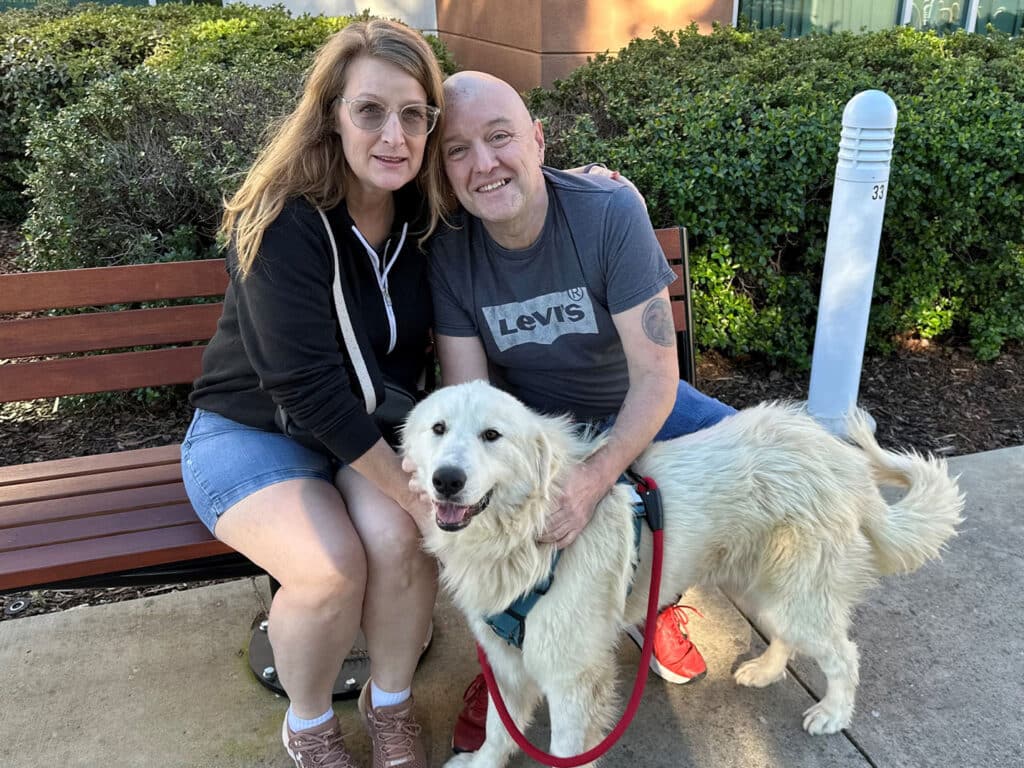 A couple sitting on a bench outside, smiling while posing with a fluffy golden retriever. The woman is wearing glasses and a black hoodie, while the man is in a gray t-shirt. The dog, leashed, appears friendly and relaxed.