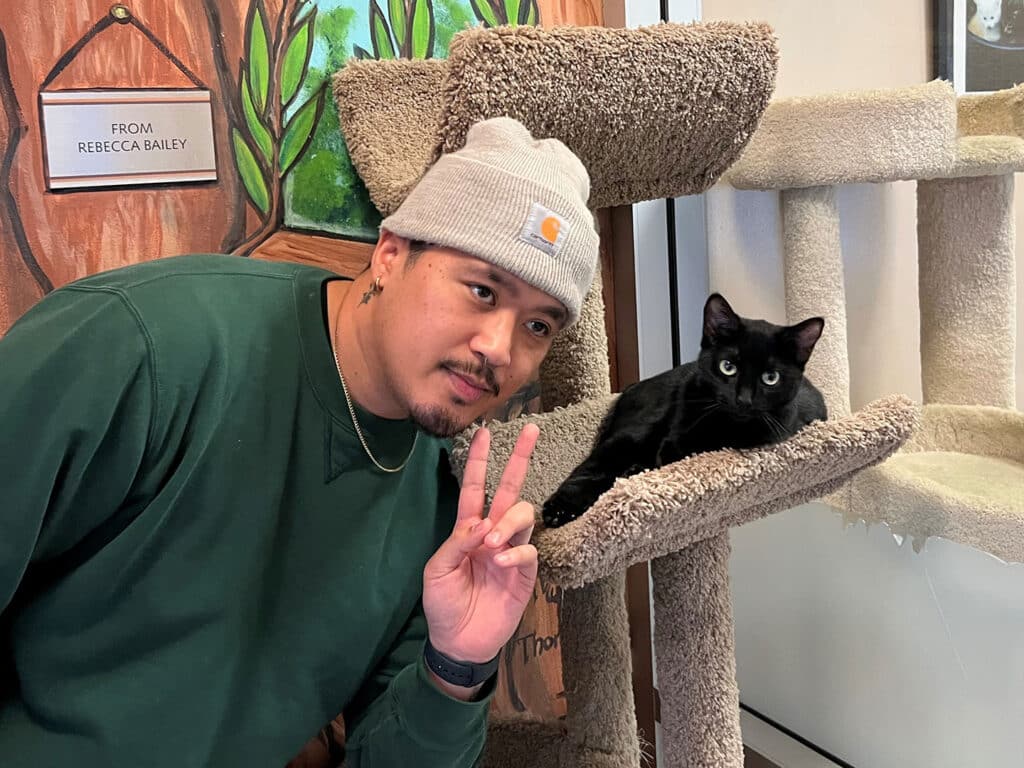 A man wearing a green sweatshirt and gray beanie poses with a peace sign next to a black cat resting on a beige cat tree. The background features a mural of trees, reinforcing the adoption theme in the setting.