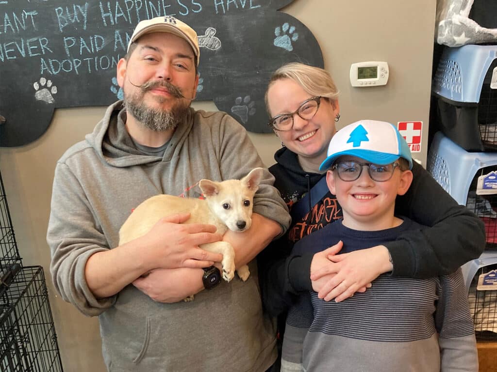 A family of four poses together, smiling while holding a small tan dog in a pet adoption center. The adults are wearing casual clothes, and the young boy is in a blue cap. The background includes pet carriers and a chalkboard with an adoption message.