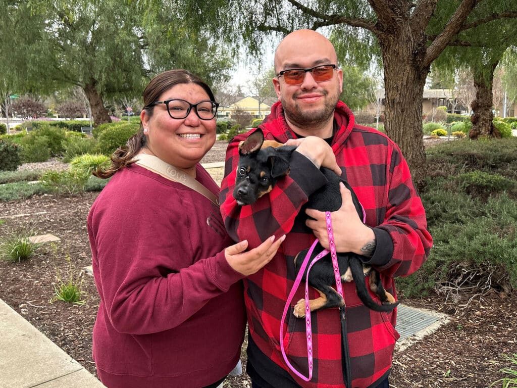 A man and a woman standing outdoors, smiling while holding a small black dog. The woman wears glasses and a maroon sweater, while the man is in a red and black checked hoodie. Greenery and landscaping are visible in the background.