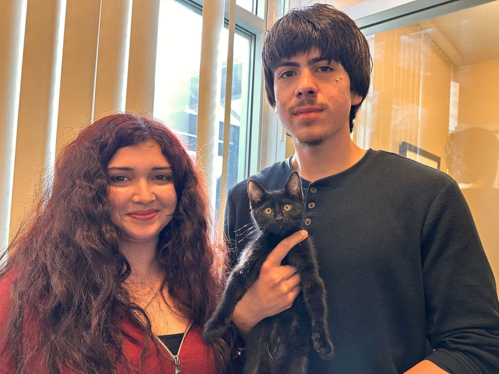 A young woman with long curly hair and a young man with short hair are standing in front of a window. The woman is smiling, holding a black kitten in her arms. The young man stands beside her with a neutral expression. Both are in an indoor setting.