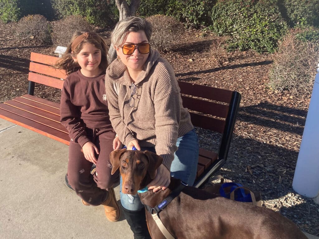 A woman and a young girl sit on a park bench with a brown dog beside them. The woman wears sunglasses and a cozy sweater, while the girl is dressed in brown attire. The scene suggests a joyful moment, possibly related to the adoption of the dog.