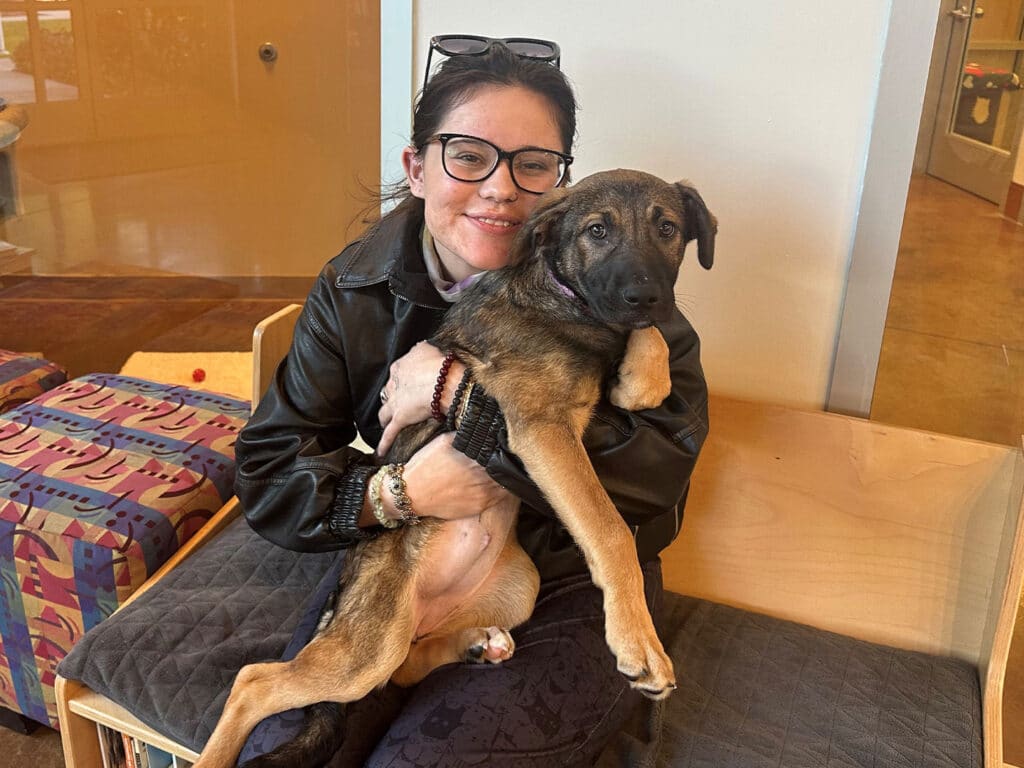 A woman with black glasses is sitting on a bench, holding a medium-sized dog close to her. The dog, with brown fur and a playful expression, is cradled in her arms. The setting appears to be a pet adoption environment.