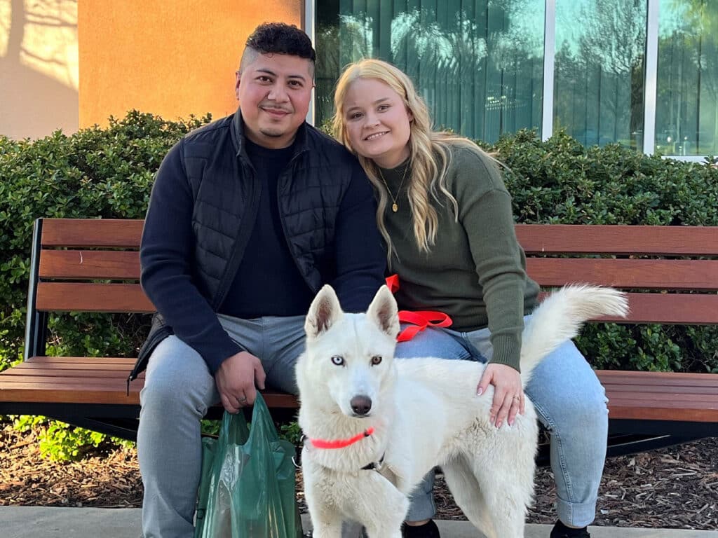 A man and a woman sitting on a bench outside, smiling while posing with a white dog wearing a red collar. The background features greenery and a building. This image represents recent adoptions, emphasizing the joy of bringing a pet into a home.