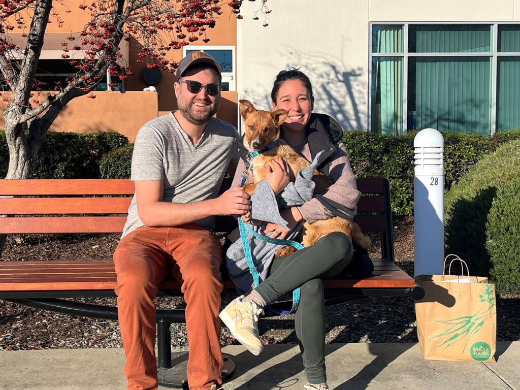 A man and a woman sit on a bench holding a small dog. The man wears sunglasses and a gray shirt with orange pants, while the woman is dressed in a gray hoodie and black leggings. A paper bag rests on the ground beside them. The setting includes colorful trees and a modern building.