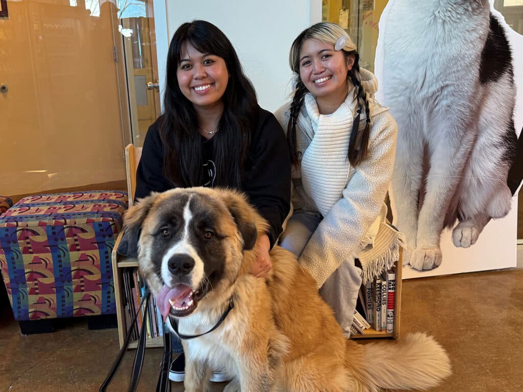 Two women sit beside a large, friendly dog, smiling at the camera. The dog, a tan and white fluffy breed, sits in front of them. The background shows a colorful patterned bench and a cutout of a dog on the wall, emphasizing themes of adoption.