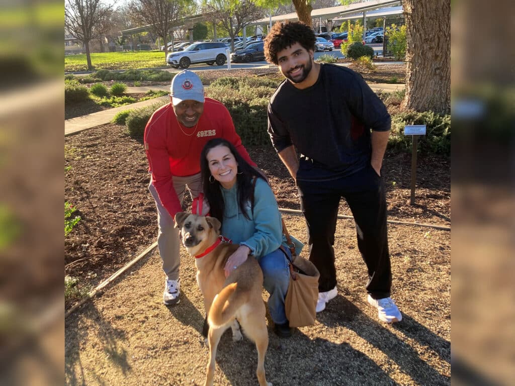 A man in a red shirt, a woman in a teal sweater, and a man in a black shirt pose together in a park, kneeling beside a medium-sized tan dog wearing a red collar. The background includes grass and shrubbery.