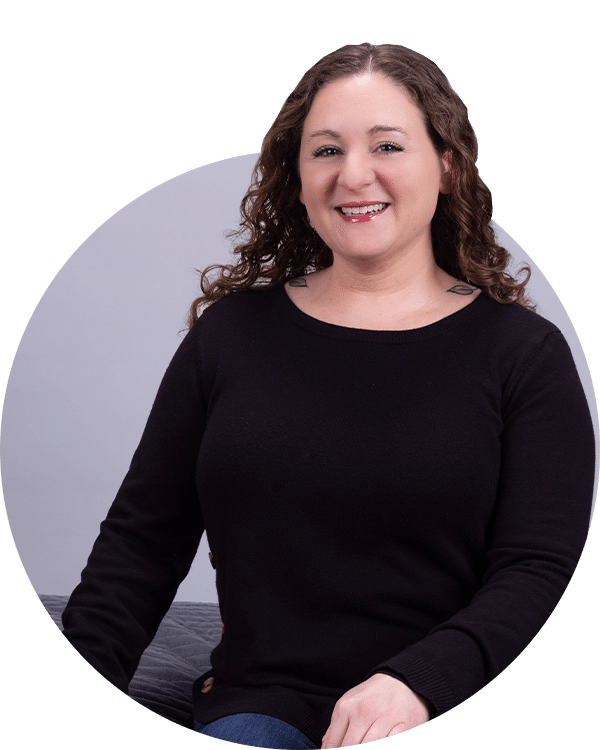 A woman with curly hair wearing a black sweater and blue jeans, sitting on a gray surface. She is smiling and posing for the camera. This image is part of the Joybound Team section, showcasing team members.