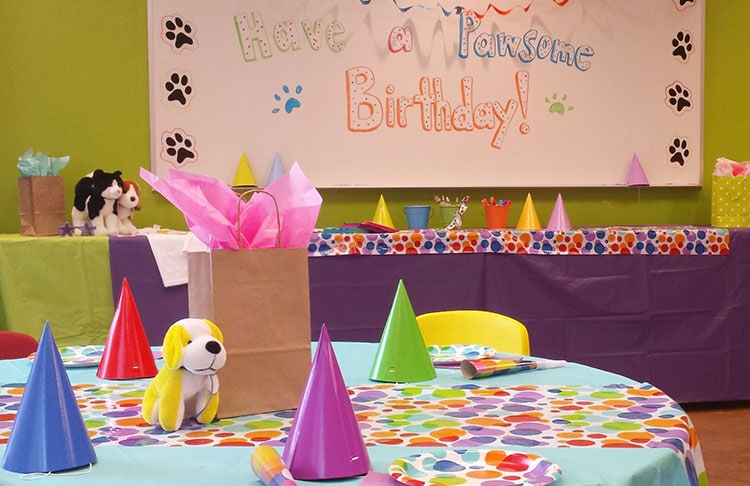A colorful birthday party setup featuring a round table with a polka dot tablecloth, party hats in various colors, and a stuffed animal. A banner on the wall reads "Have a Pawsome Birthday!" with gifts and decorations in the background.