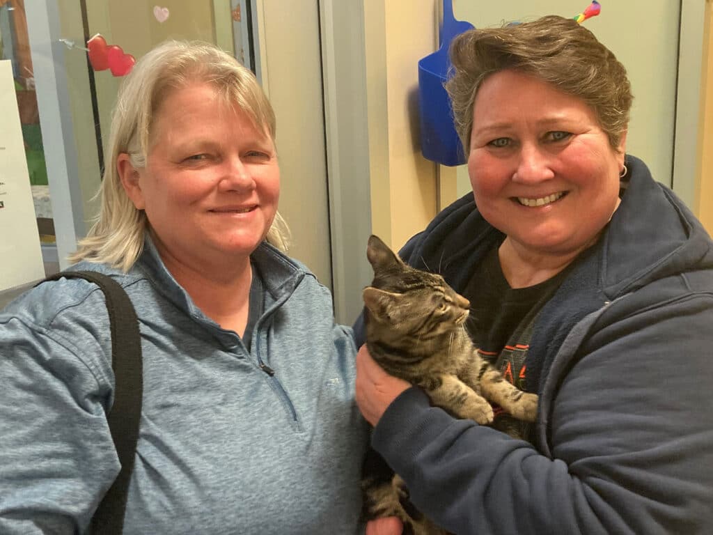 Two women smiling while holding a gray tabby cat in a room with light-colored walls and a window. The image represents a recent adoption, showcasing the joy of bringing a pet into a new home.