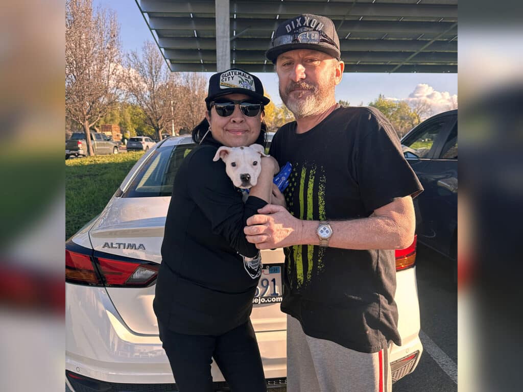 A couple standing outside near a white car, holding a small puppy. The woman wears a black hoodie and baseball cap, while the man is in a black T-shirt and cap. They smile, appearing happy after their recent adoption of the puppy.