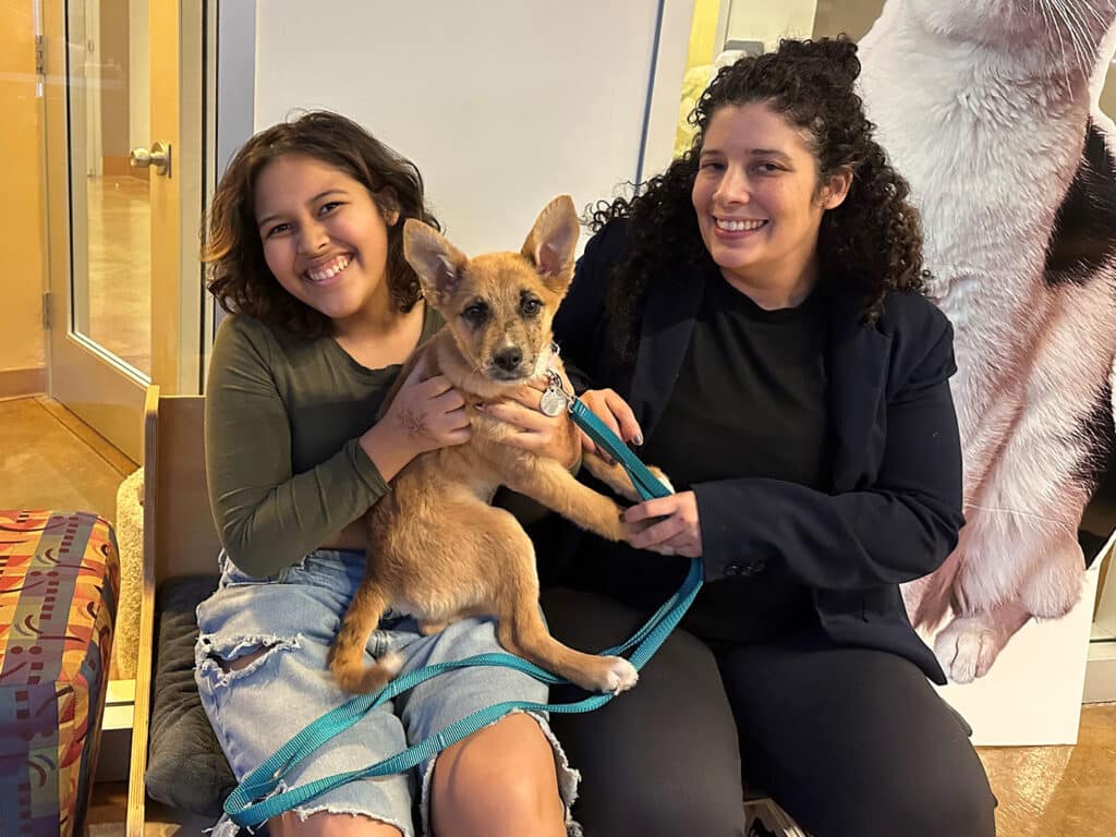 Two women sitting with a brown puppy on a leash in a welcoming environment. One woman is smiling while holding the puppy, and the other is sitting beside her, also smiling. The background features a large cutout of a dog.
