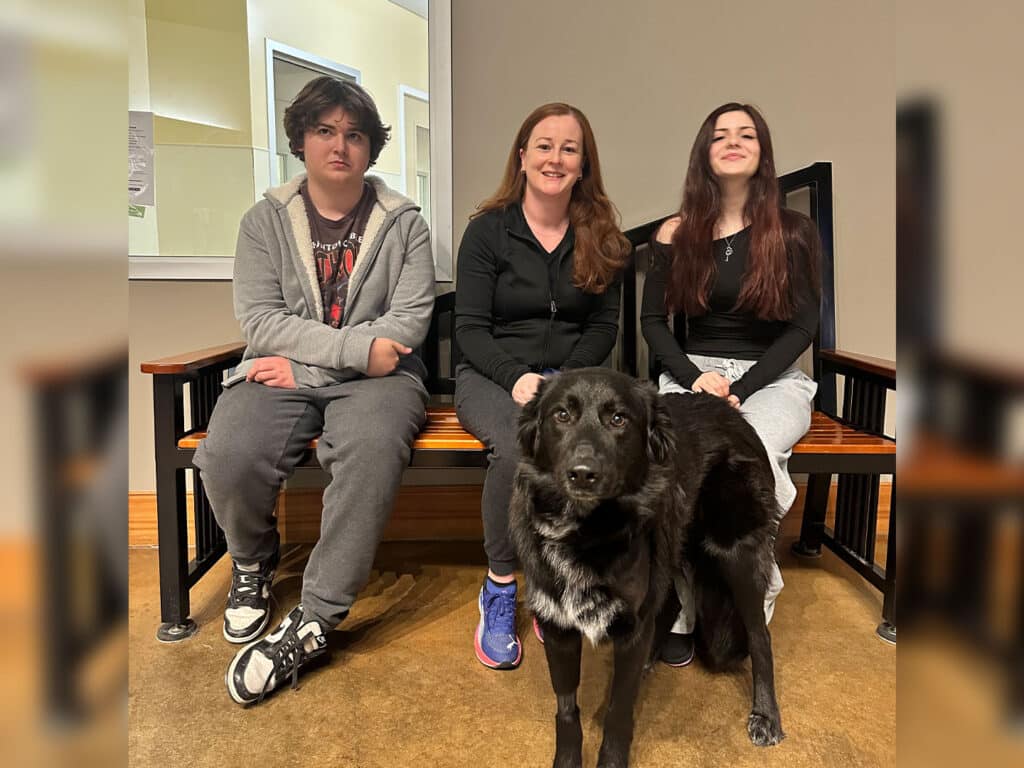A family sitting on a bench in an adoption center, featuring a young boy, a woman, and a young girl, with a black dog in front of them. The setting is indoors with a light background, and the family is smiling while the dog appears calm.