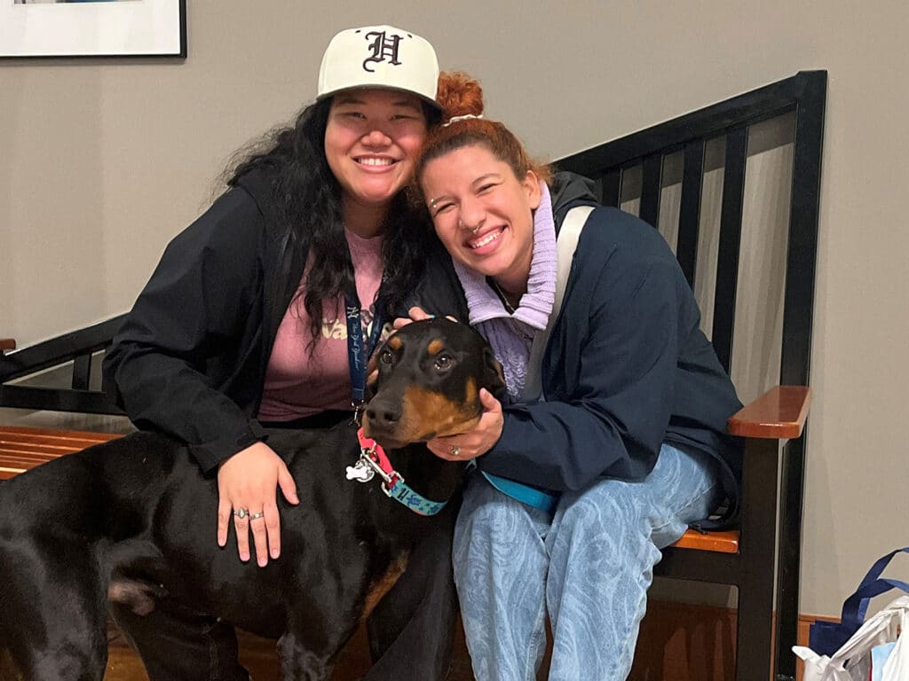 Two women sitting on a bench smiling with a black dog, who has a blue collar. One woman wears a white cap and the other has curly hair, both are casually dressed. The setting is a cozy indoor space, indicating a moment of joy possibly related to pet adoption.