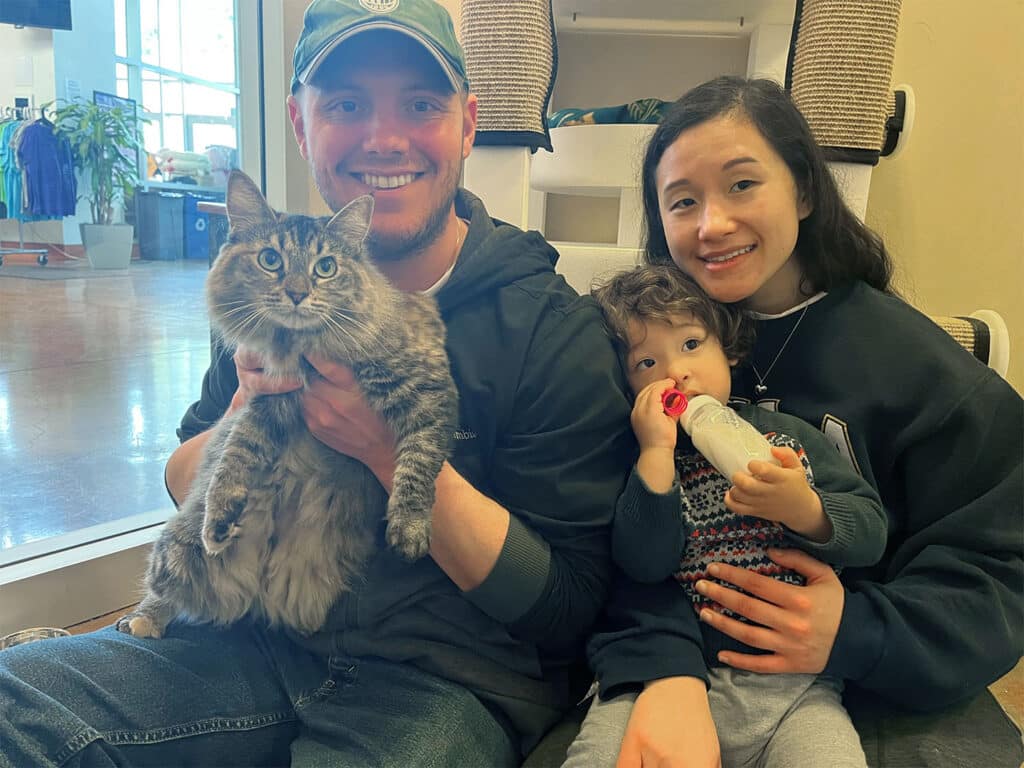 A family of three is sitting together in a bright room. A man holds a fluffy gray cat in his arms, while a woman sits beside him, holding a young child who is drinking from a bottle. The atmosphere suggests a focus on adoption and companionship.