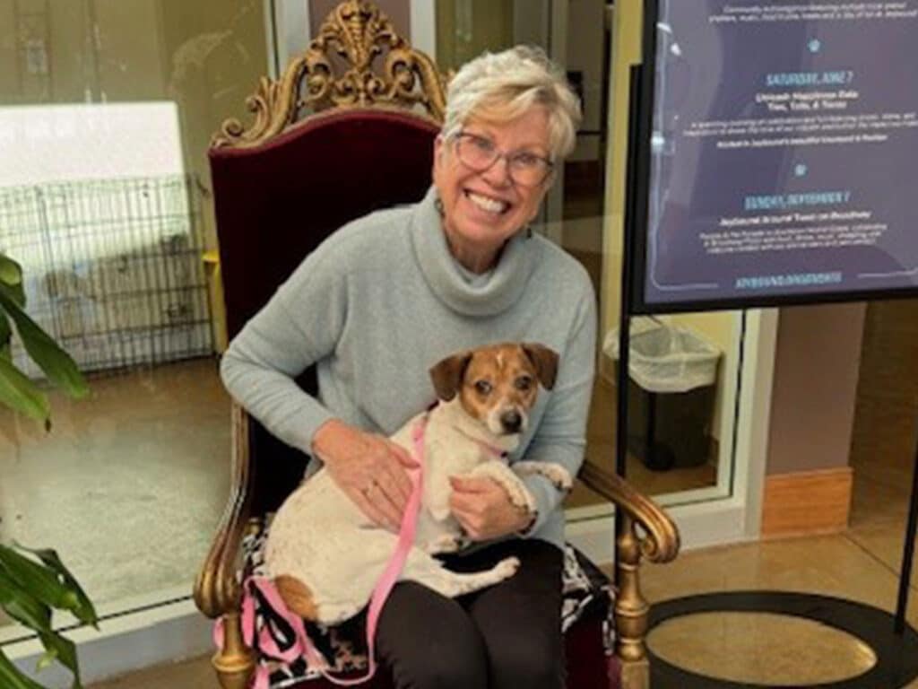 A woman with short gray hair smiles while sitting on an ornate chair, holding a small dog on her lap. The dog has brown and white fur. A sign is visible in the background, promoting adoption events.
