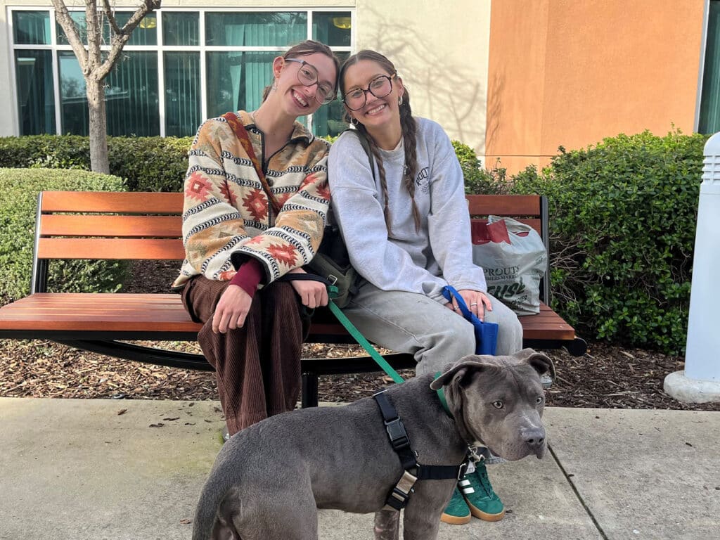 Two young women sitting on a bench outside, smiling at the camera. One is wearing a patterned sweater and the other in a gray sweatshirt. A gray dog on a leash sits between them. The background features greenery and a building, relating to an adoption theme.