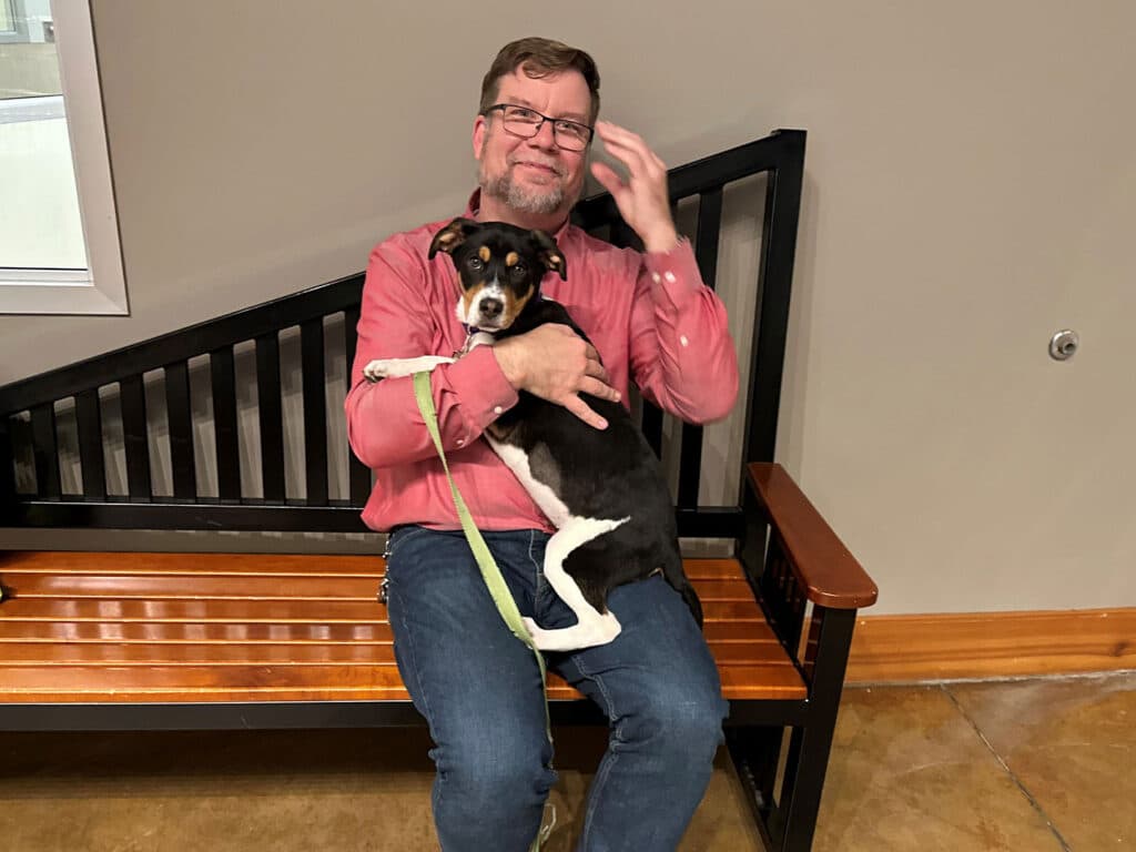 A man sitting on a wooden bench holds a small dog in his lap. The man wears glasses and a red shirt, smiling gently. The dog, with black and white fur, appears relaxed and comfortable in his embrace. This image emphasizes the theme of Adoption.