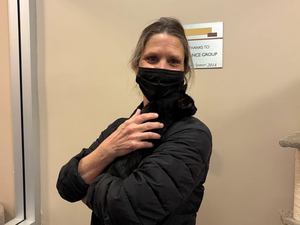 A woman wearing a black mask holds a small black cat close to her chest. She has long hair and is dressed in a black jacket. The background shows a wall with a plaque, indicating a community group, with warm lighting enhancing the scene. This image relates to cat adoption.