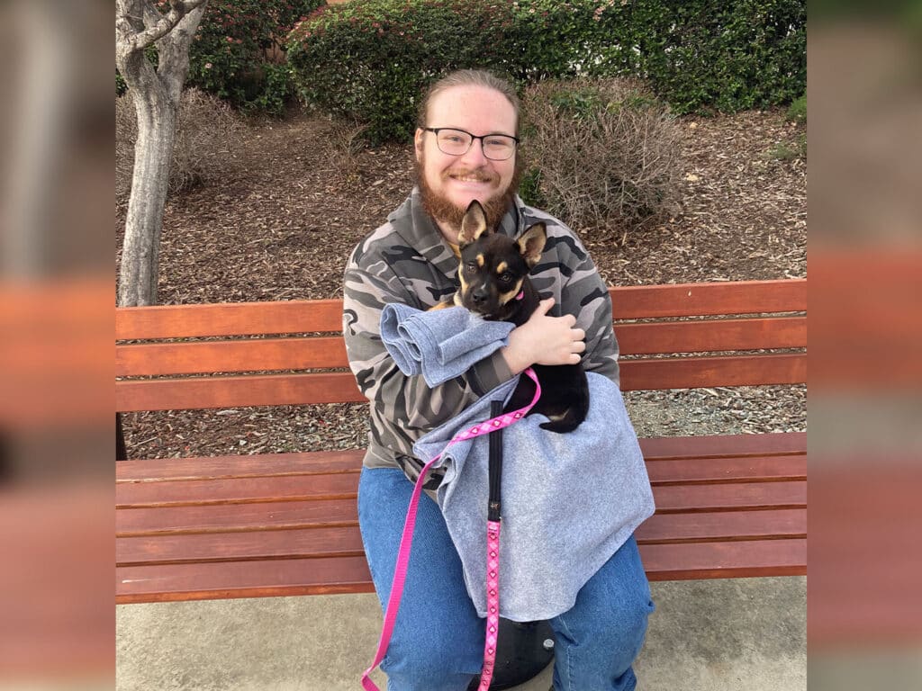 A man with glasses smiling while sitting on a wooden bench, holding a small dog wrapped in a gray blanket. The dog is black with tan markings. The surrounding area features greenery and bushes, providing a natural backdrop.