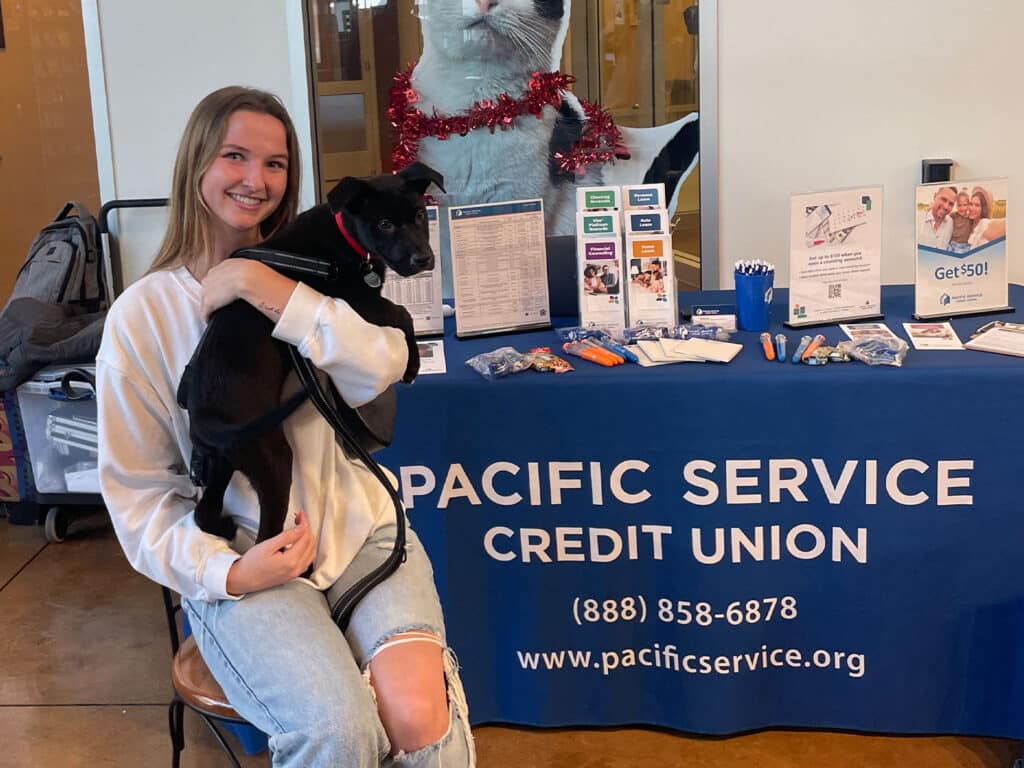A young woman holding a small dog while sitting on a stool in front of a blue table displaying information about Pacific Service Credit Union. Adoption resources are visible among the materials on the table.