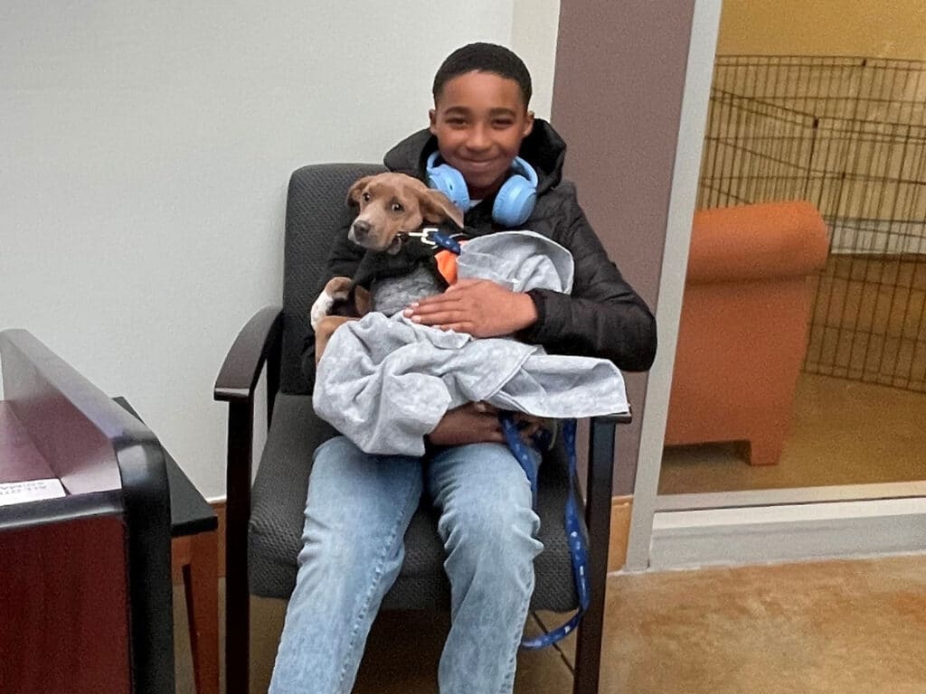 A young boy sitting in a chair, holding a small dog wrapped in a grey blanket. The boy is smiling and wearing headphones. The setting appears to be an animal shelter or adoption center, indicating a recent adoption.