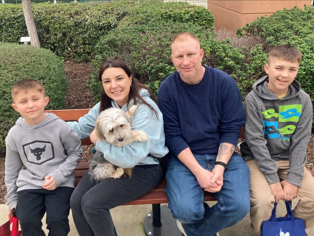 A family sitting on a bench outside, consisting of two boys, a girl holding a small dog, and an adult man. They are all smiling, with greenery in the background, suggesting a moment of joy related to adoption.