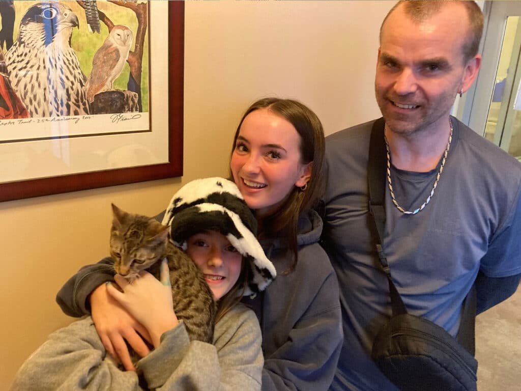 A family gathers in a hallway, smiling together while holding a cat. Two children, one wearing a cow-patterned hat, are smiling and gently holding the tabby cat. An adult man stands beside them, also smiling. A colorful bird illustration is visible on the wall.