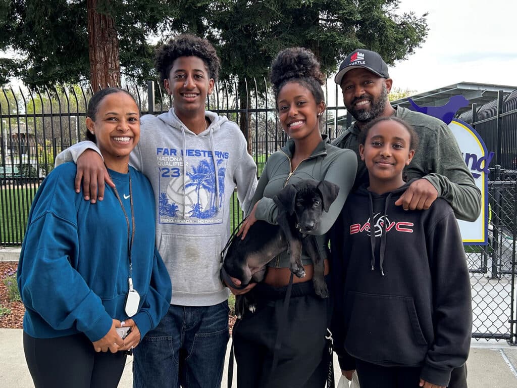 A family stands together outdoors, smiling for the camera. The group includes a woman on the left, three children, and a man holding a puppy. The setting appears to be a park, suggesting a joyful moment related to pet adoption.
