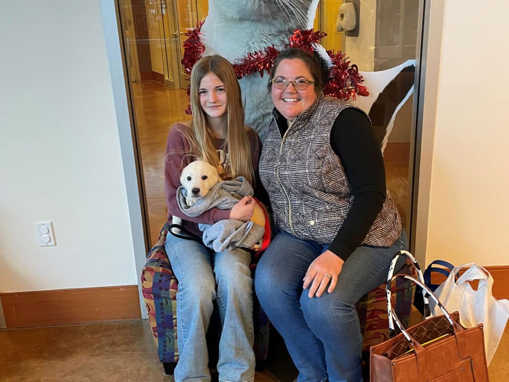 A young girl holds a small puppy wrapped in a blanket while sitting next to a smiling woman. They are seated on a patterned chair, with a festive decoration in the background. The image highlights a recent moment of adoption.