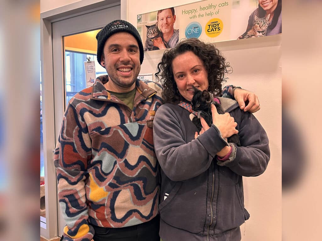 A man and a woman stand together inside an animal adoption center, smiling at the camera. The woman holds a small black kitten. Colorful banners in the background promote happy, healthy cats and support for adoption.