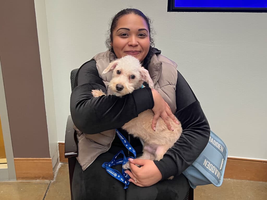 A woman sitting in an office chair holds a small white dog in her arms, smiling gently. The setting suggests a recent adoption, with the dog wearing a blue leash. The woman is dressed in a black outfit with a vest.