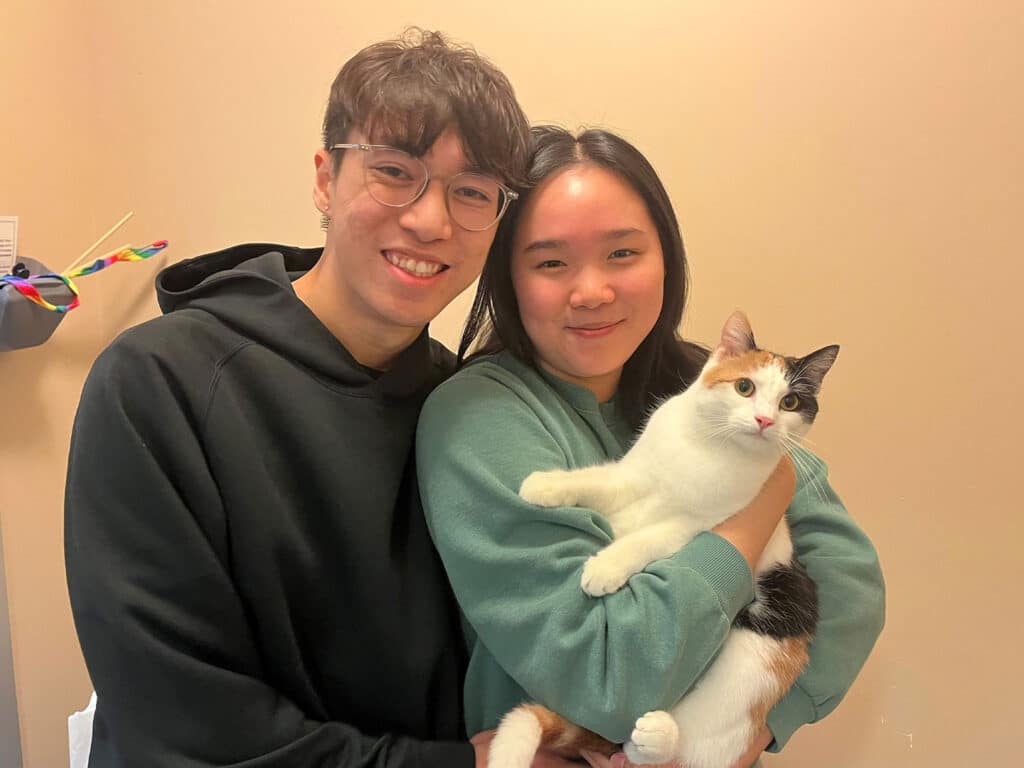 A young man and woman stand together, smiling while holding a calico cat in a cozy room. The woman wears a teal sweater, and the man wears a black hoodie. This image highlights their recent adoption of the cat, emphasizing the joy of adopting pets.