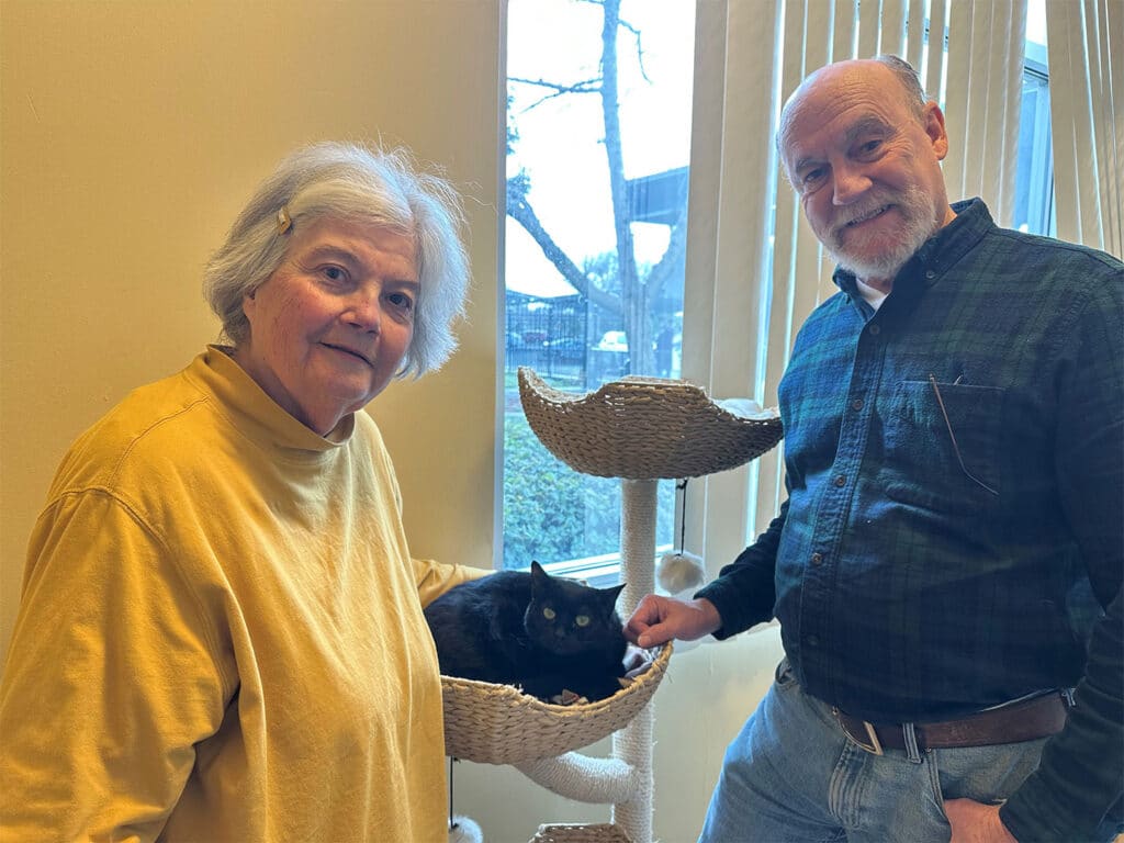 An older woman and man stand beside a cat scratching post, with a black cat resting inside a basket. They smile towards the camera, in a cozy room with natural light coming through a window. This image celebrates the theme of adoption.