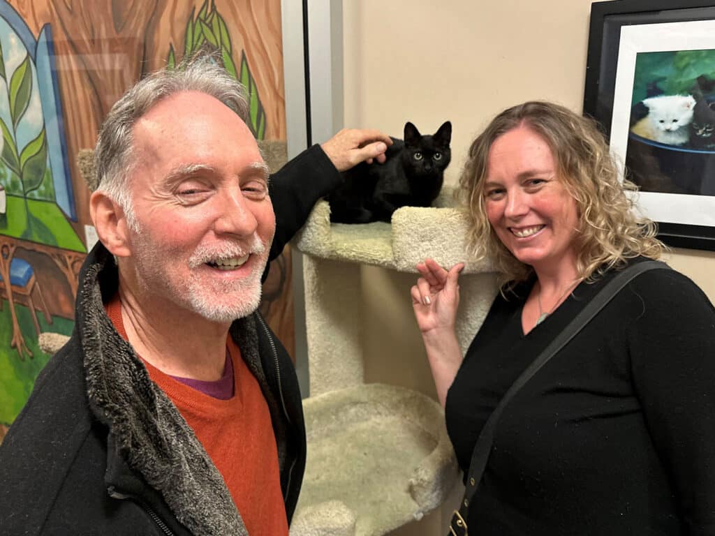 A man and a woman smile while standing next to a cat tree. A black cat rests on a level of the cat tree. The background features a colorful mural of animals. This image relates to an adoption event and the joy of interaction with pets.