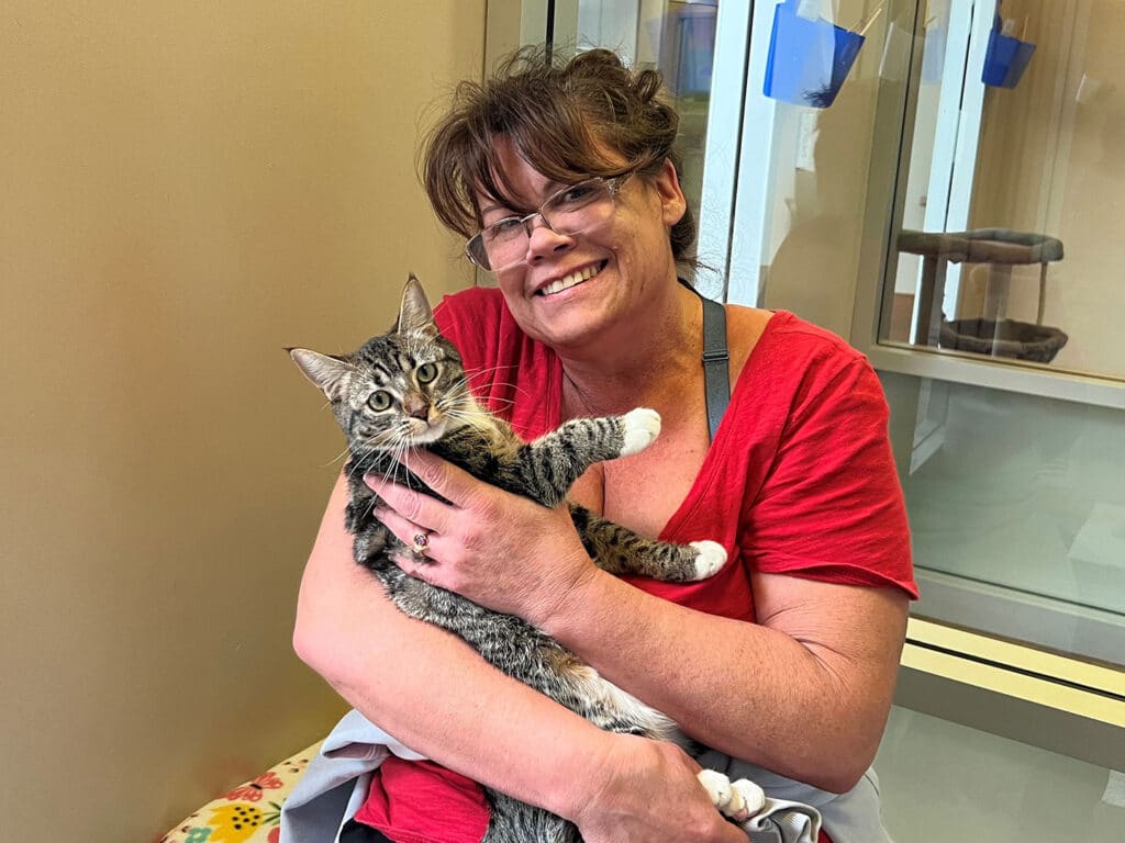 A woman in a red shirt smiles while holding a tabby cat in her arms at an animal shelter. The background features a glass enclosure and various pet supplies. The image highlights the theme of Adoption.