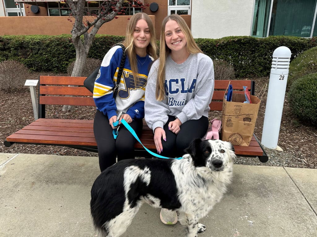 Two young women sitting on a bench outside, smiling while holding a dog on a leash. The dog is black and white with a fluffy coat. In the background, there are plants and part of a building visible, suggesting a community or park setting related to adoption.