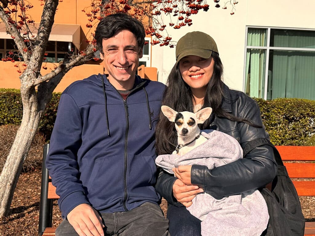 A smiling couple sits on a bench outdoors, with a small dog wrapped in a blanket held by the woman. The scene features a tree with red leaves and a modern building in the background. The image highlights themes of adoption and companionship.