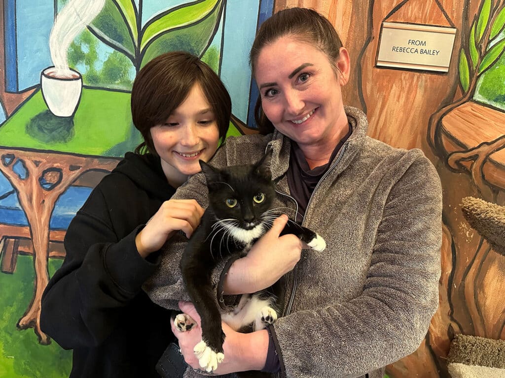 A woman and a child are smiling while holding a black and white cat in a colorful room with plants and a coffee cup mural. The image highlights a moment of joy related to cat adoption.