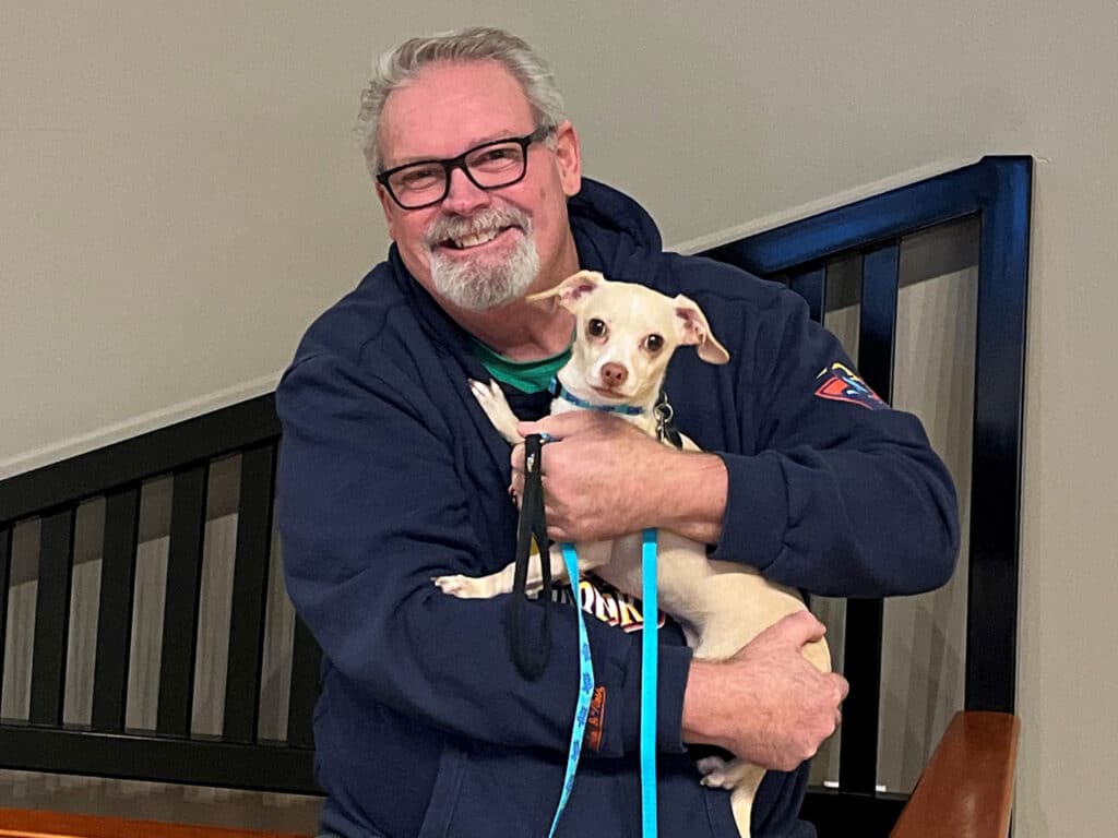 A man with glasses smiling while holding a small beige dog in his arms. The background shows a staircase and a wall. This image highlights the theme of adoption.
