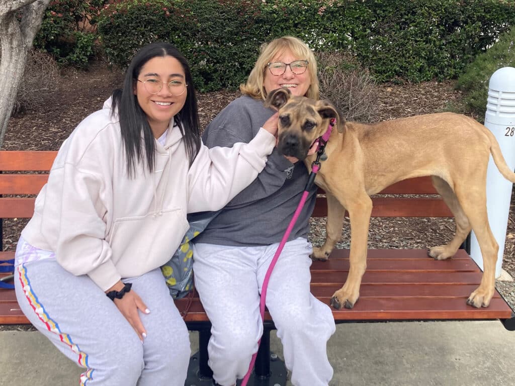 A young woman and an older woman sitting on a park bench, both smiling. The young woman is gently holding a dog on a leash, while the dog stands beside her with its front paw on the older woman's lap. Adoption theme apparent.