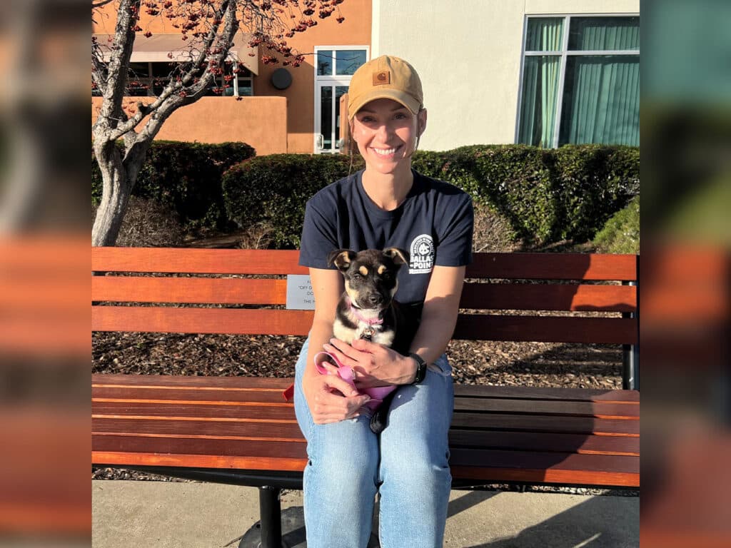 A smiling woman wearing a blue shirt and a beige cap sits on a bench, holding a small dog in her lap. The dog wears a pink harness. Behind them is a backdrop of bushes and buildings, with sunlight casting shadows.