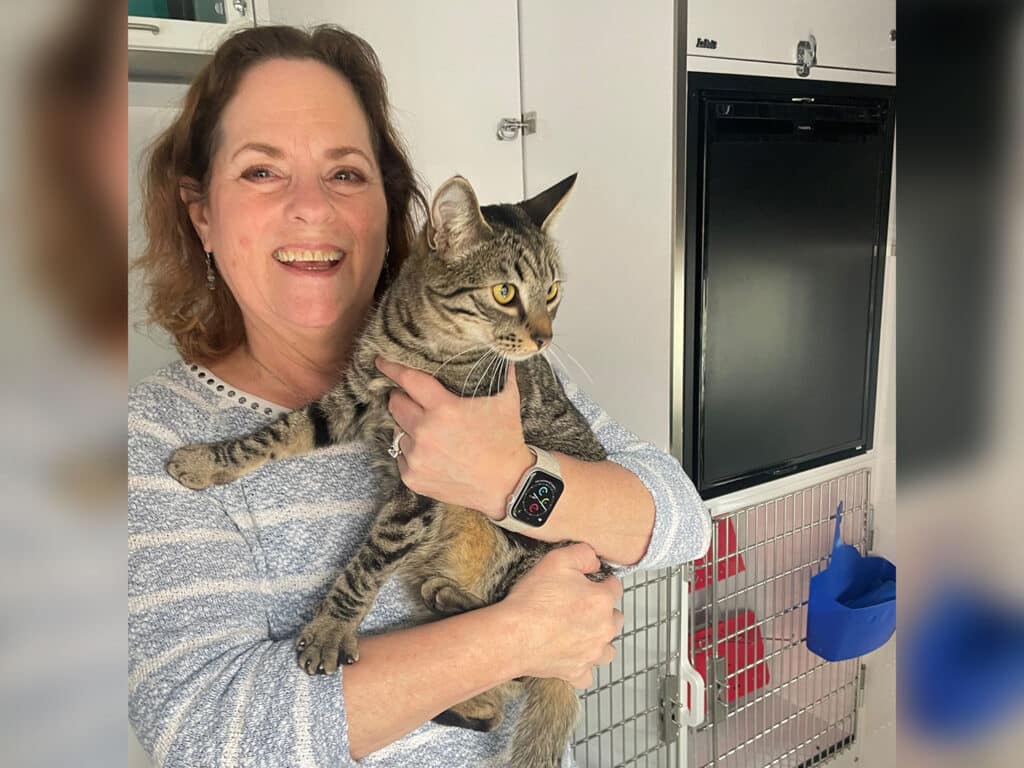 A woman smiling while holding a gray tabby cat in a pet adoption center. She wears a striped sweater and has a smartwatch on her wrist. Behind them is a metal cage with pet supplies, highlighting the adoption theme.