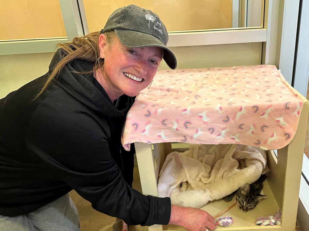 A woman wearing a black hoodie and a cap smiles while gently reaching into a small pet enclosure. Inside, a tabby cat is nestled within a warm blanket. A pink patterned cover drapes over the enclosure. The image relates to adoption.
