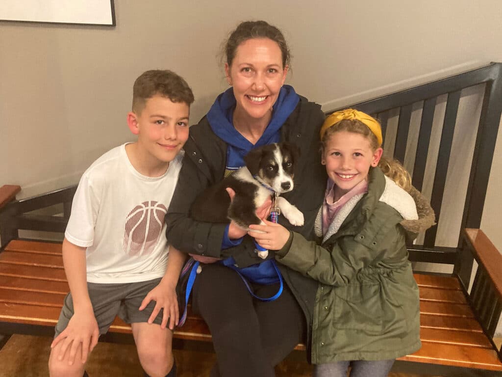 A woman sits on a bench holding a small black and white puppy, flanked by two children. The boy on the left wears a white T-shirt and gray shorts, while the girl on the right is in a green jacket and scarf. The setting appears welcoming, hinting at a recent dog adoption.