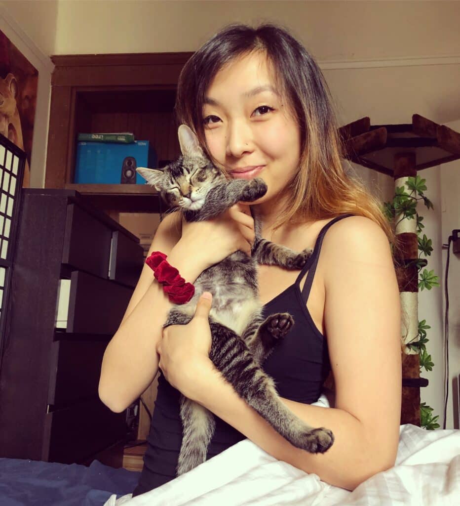 A woman with long hair holds a tabby cat in her arms, smiling. She is wearing a black tank top, and the background shows a cozy room with a bookshelf and a cat tree. This image reflects the companionship often associated with service animals.