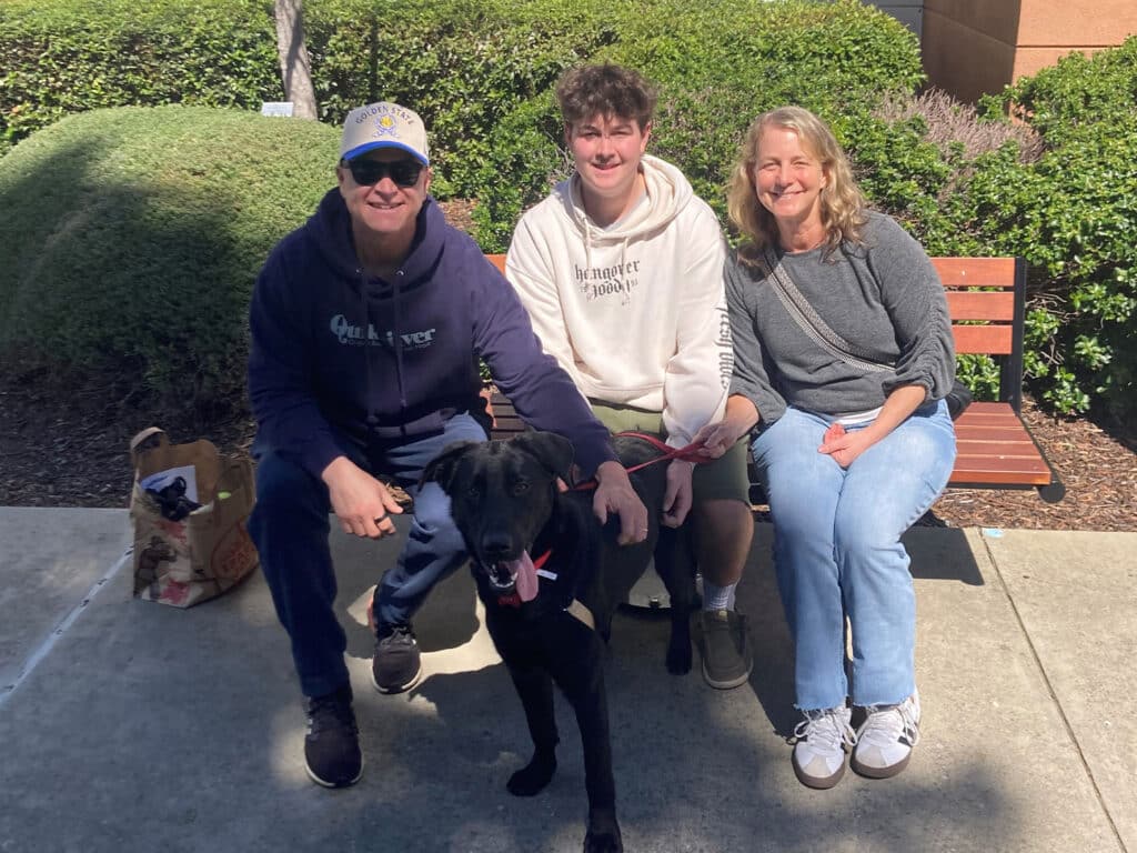 A family of three sits outdoors on a concrete surface, smiling and posing for the camera. They are accompanied by a black dog on a leash. The background features green shrubs and a bench, indicating a sunny day, possibly celebrating an adoption.